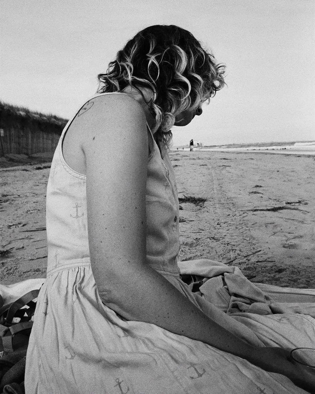 Black and white photograph of a woman with curly hair sitting on a beach, wearing a sundress with anchor prints in soft daylight.