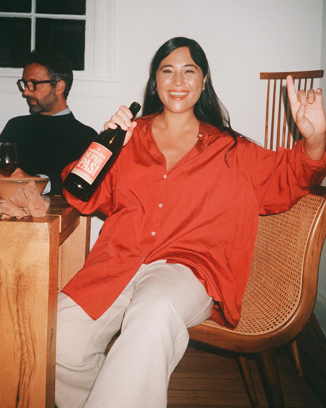 Woman in red shirt holds wine bottle indoors with friend, smiling, celebrating a special occasion or gathering for food and drinks.