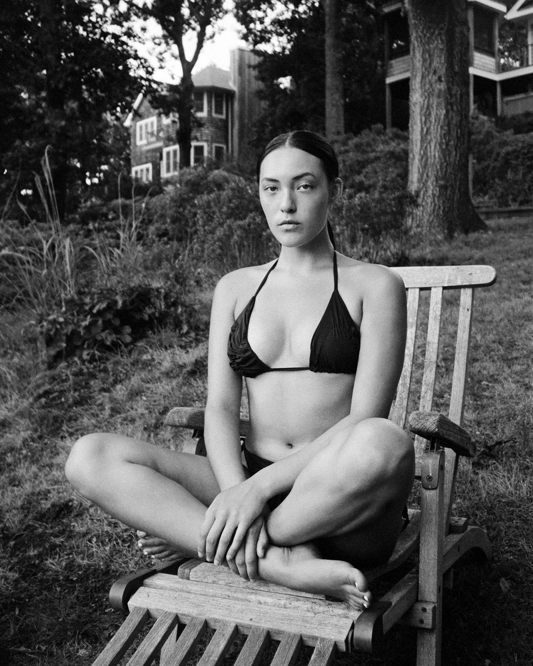 Black and white portrait of a woman in a bikini sitting on a wooden chair outdoors, with a serene expression and natural setting.