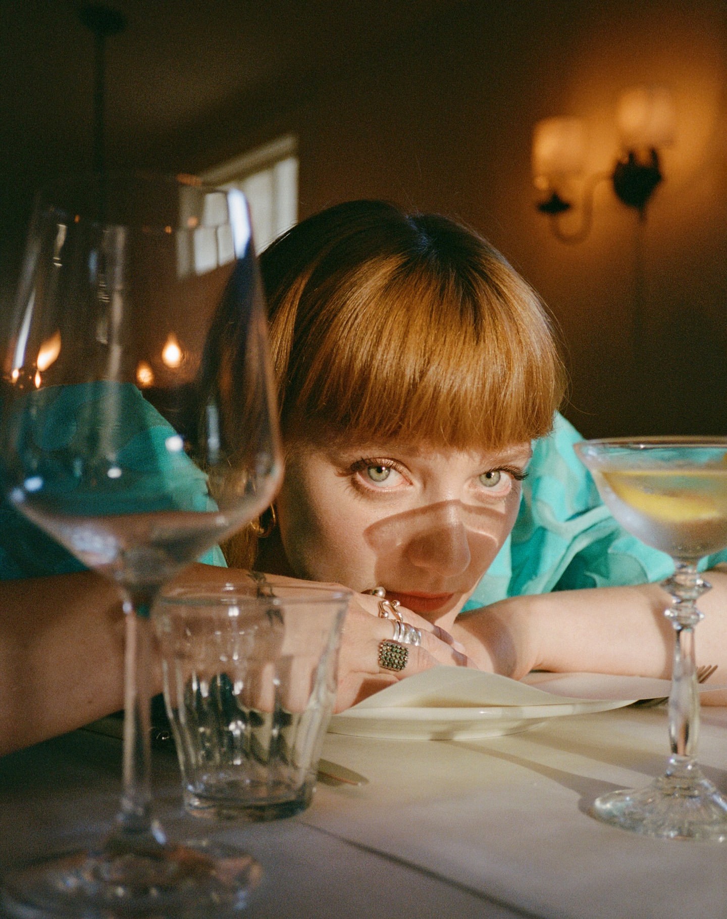 Young woman with red hair at restaurant table with cocktail and wine glasses, wearing turquoise blouse and rings looking into camera.