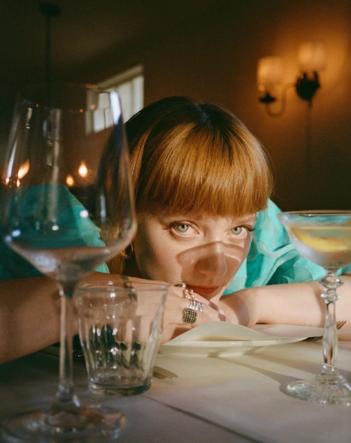 Young woman with red hair at restaurant table with cocktail and wine glasses, wearing turquoise blouse and rings looking into camera.
