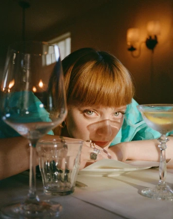 Young woman with red hair at restaurant table with cocktail and wine glasses, wearing turquoise blouse and rings looking into camera.