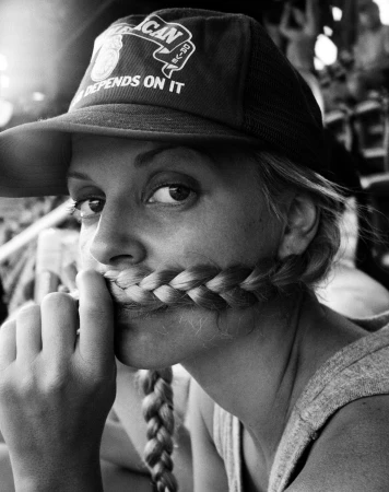 Close-up black and white portrait of a woman wearing a baseball cap and holding her braided hair across her mouth like a mustache.