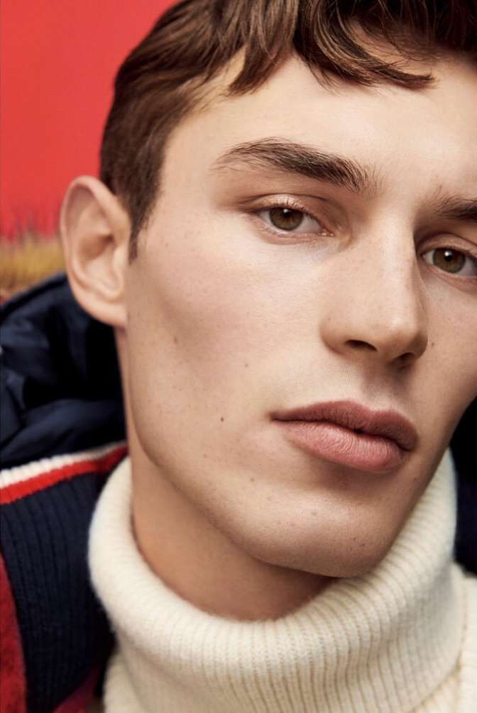 Close-up portrait of a fair-skinned adult male with brown hair and eyes, wearing a cream turtleneck sweater and a navy jacket, against a red backdrop.
