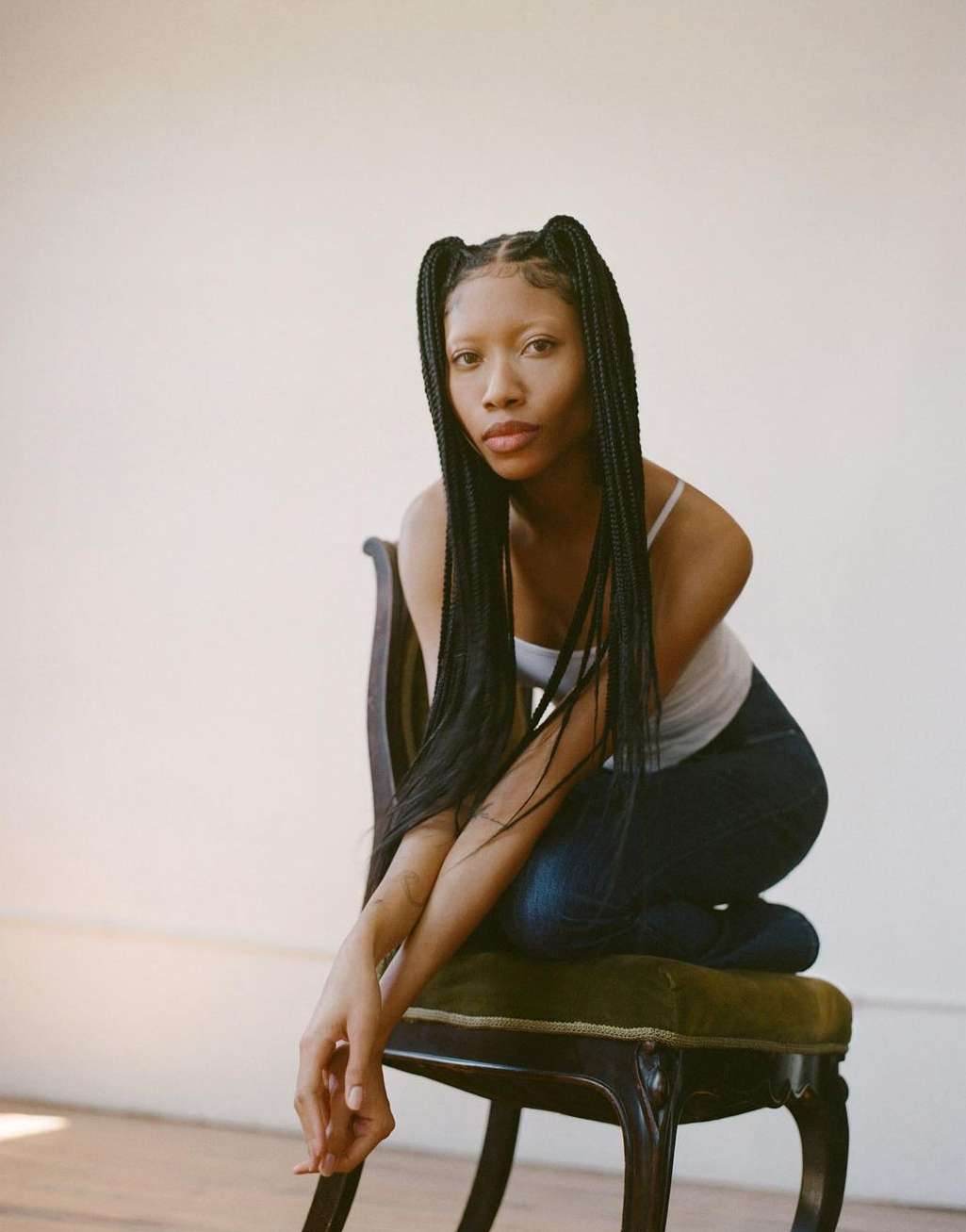 Young woman poses on a vintage chair in a light-filled studio. She wears braids, a white top, and dark jeans in a portrait shot.