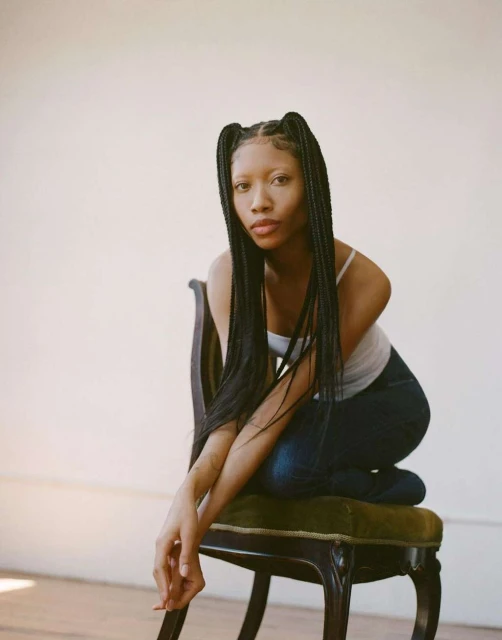 Young woman poses on a vintage chair in a light-filled studio. She wears braids, a white top, and dark jeans in a portrait shot.