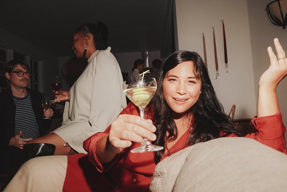 Attractive woman in a red shirt holding a martini at an indoor party with other guests in the background. Social event.