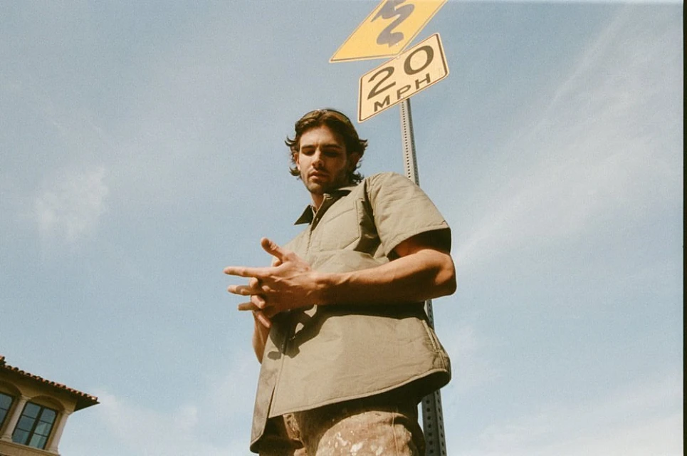 Low angle shot of a young adult man in an olive-green shirt standing by a yellow 20 MPH speed limit sign on a sunny day outdoors.