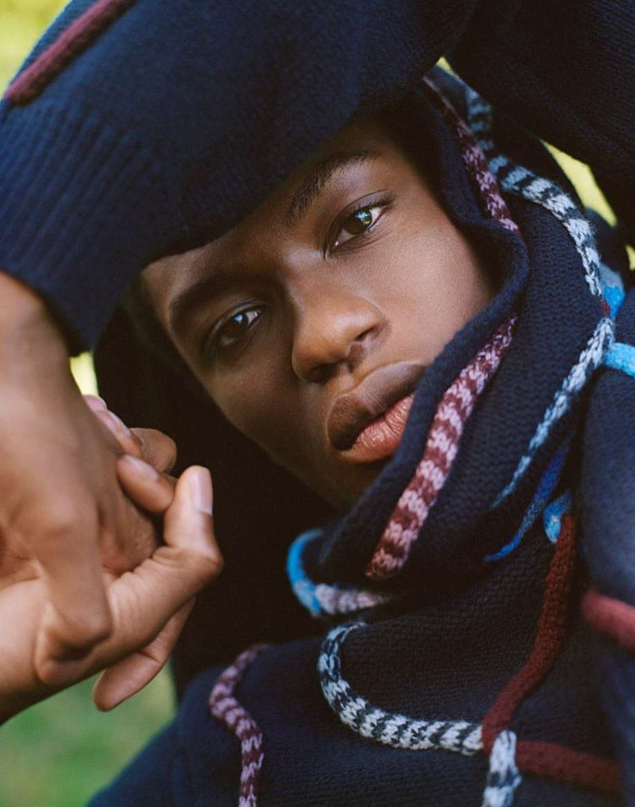 Portrait of a black male model wearing a sweater with hands near his face in an outdoor environment with green foliage.
