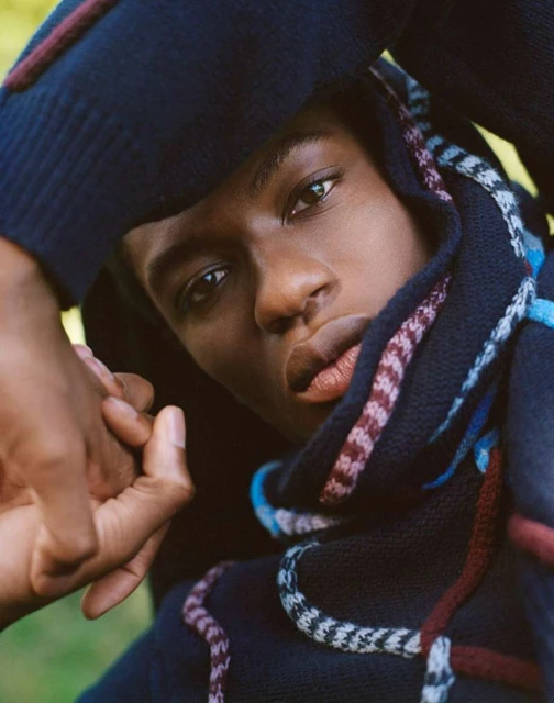Portrait of a black male model wearing a sweater with hands near his face in an outdoor environment with green foliage. 