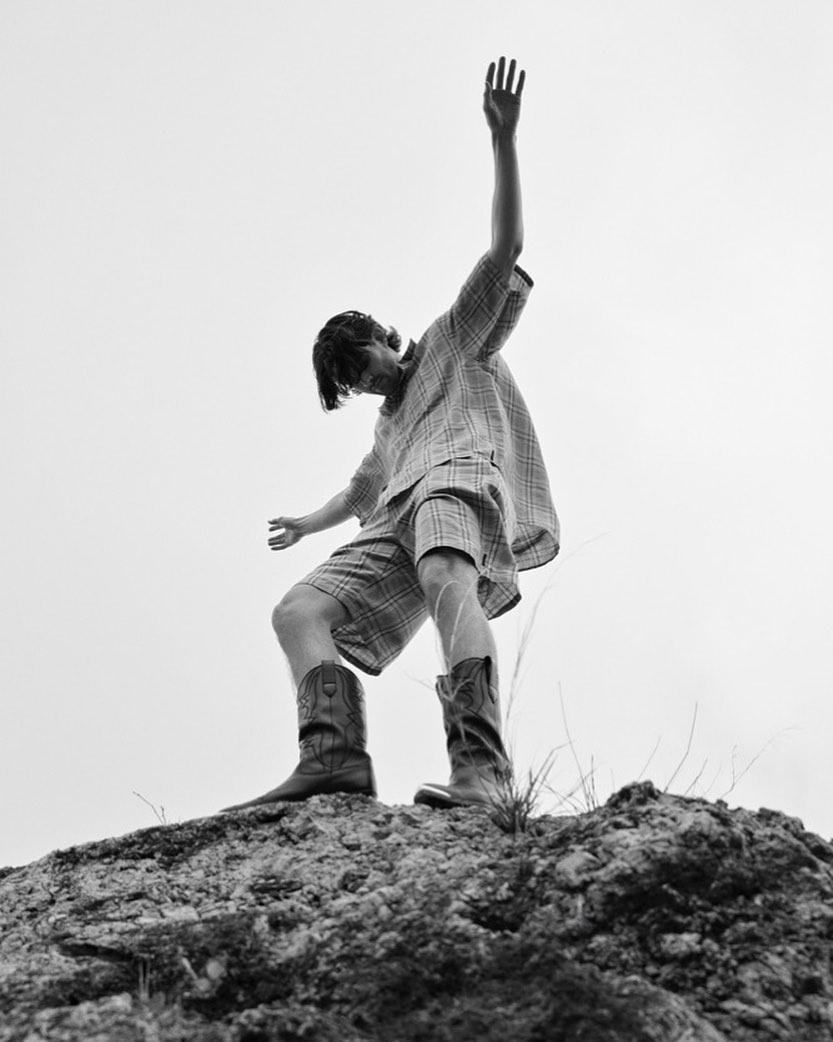 Man wearing plaid shirt and shorts with cowboy boots standing atop a rocky hill against a clear sky