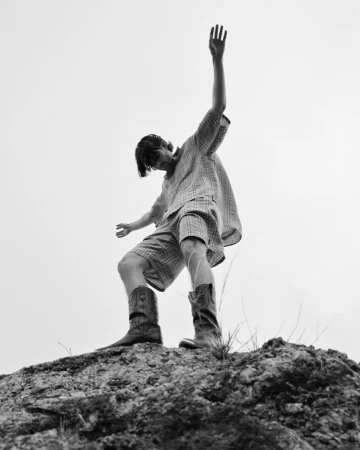 Man wearing plaid shirt and shorts with cowboy boots standing atop a rocky hill against a clear sky