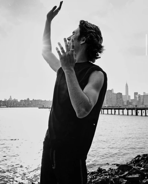 Monochrome portrait of a man with curly hair, wearing a tank top, gesturing towards the sky against a backdrop of a city skyline and water.