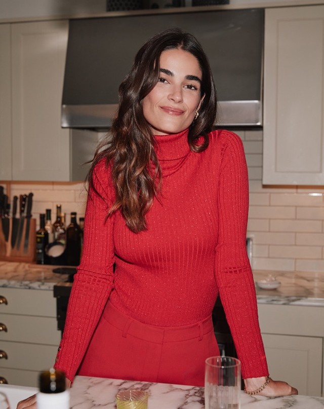 Brunette woman in red turtleneck sweater and matching pants posing in a home kitchen with neutral cabinets and marble countertops.