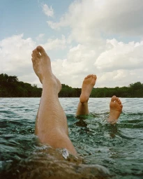 Bare legs and feet sticking up out of calm outdoor water with distant treeline and cloudy sky under natural light