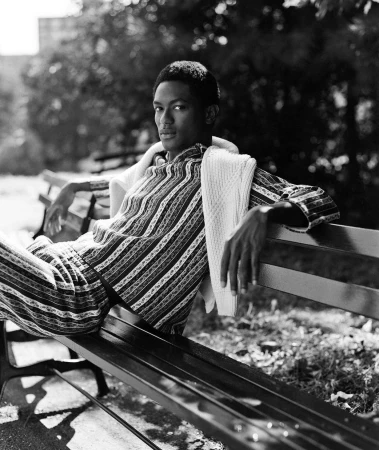 Monochrome portrait of a young man wearing patterned shirt and trousers, seated on an outdoor park bench.