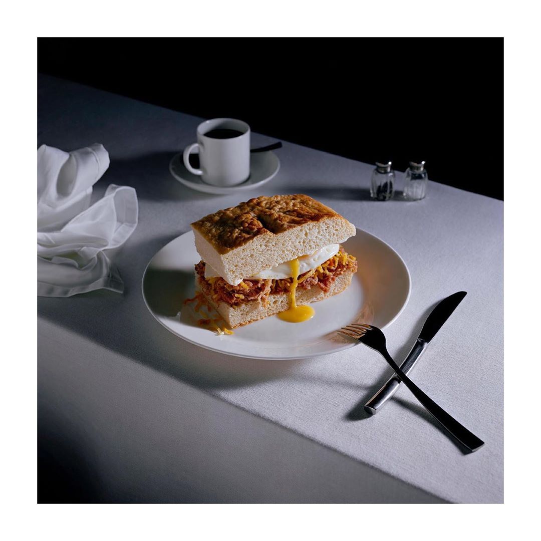 Elevated shot of a sandwich with a dripping fried egg, a cup of coffee, and cutlery on a white tablecloth against a dark background.