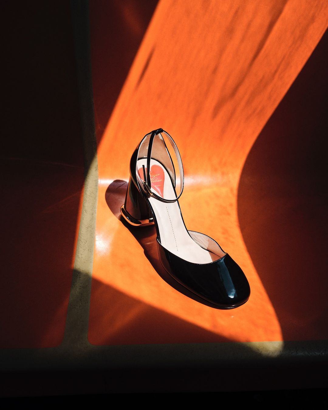 High-angle shot of a shiny black heeled sandal with an ankle strap on an orange background, casting interesting light patterns.