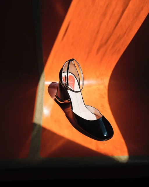 High-angle shot of a shiny black heeled sandal with an ankle strap on an orange background, casting interesting light patterns.