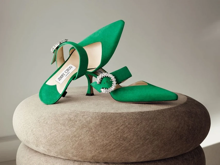 Emerald green Jimmy Choo heels with crystal buckle detail, displayed on a textured beige surface in a brightly lit studio.