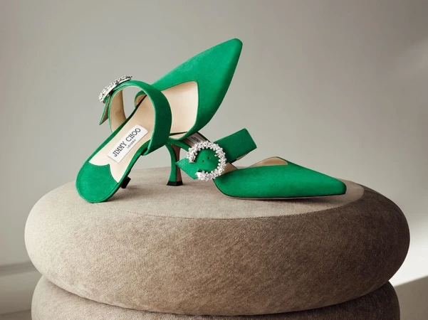 Emerald green Jimmy Choo heels with crystal buckle detail, displayed on a textured beige surface in a brightly lit studio.