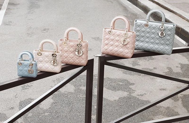 Group of five light blue and pink Lady Dior handbags in varying sizes displayed on a black railing against a grey surface.