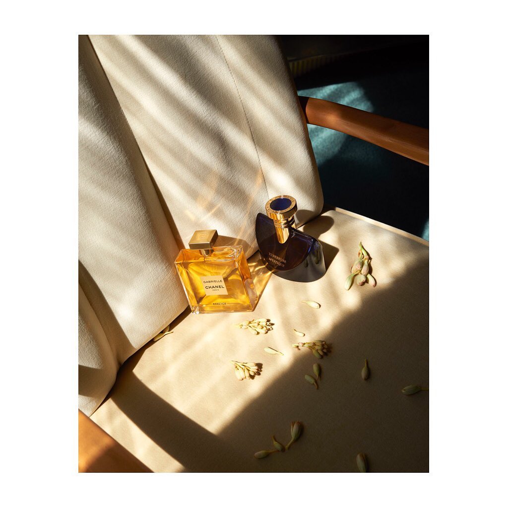 Still life composition featuring two luxury perfume bottles, small white blossoms, and a draped cream-colored blazer on a wooden chair.