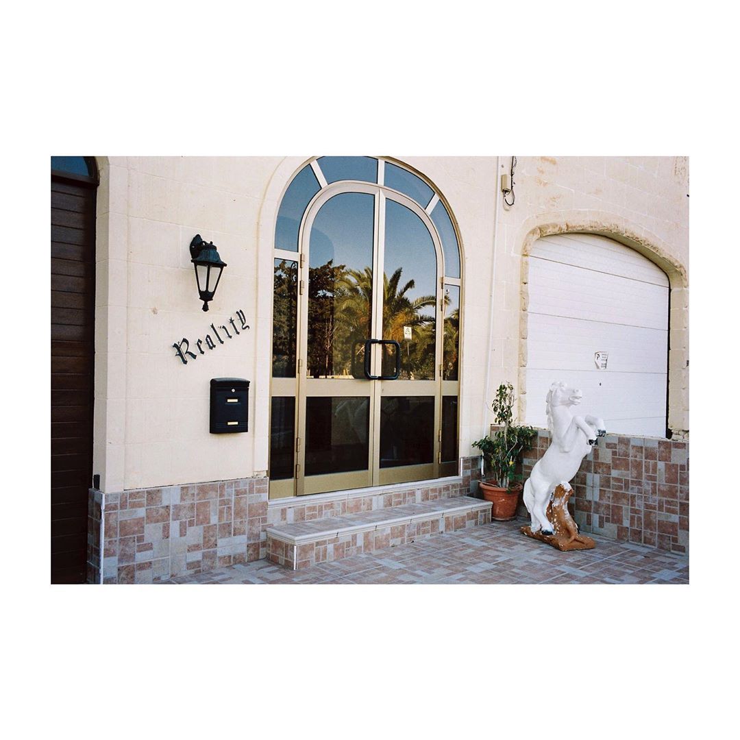 Architectural shot of a light beige building with a glass door, a wrought-iron lamp, and a white horse sculpture near the entrance.