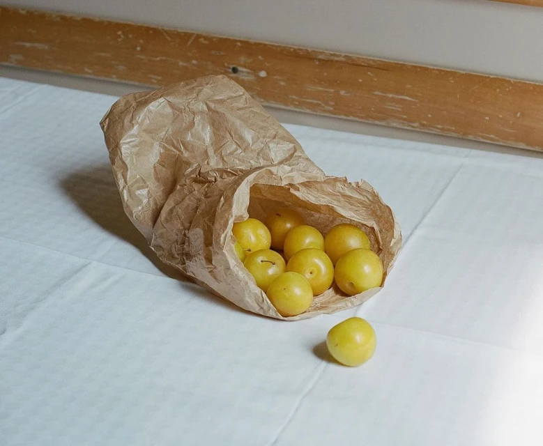 Close-up of yellow plums spilling out of a crumpled brown paper bag on a white surface, creating a simple food still life.