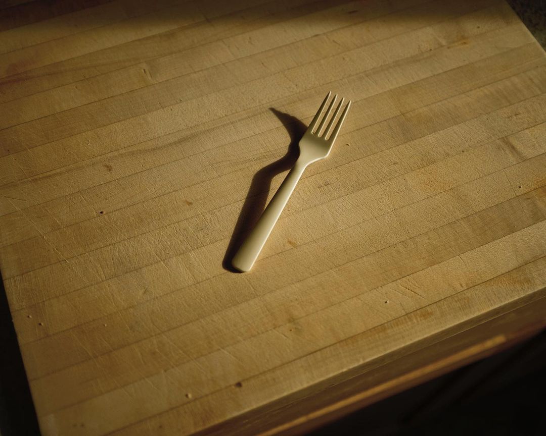 Close-up of a beige fork on a wooden surface, illuminated by warm lighting, casting distinct shadows and highlighting the wood grain.