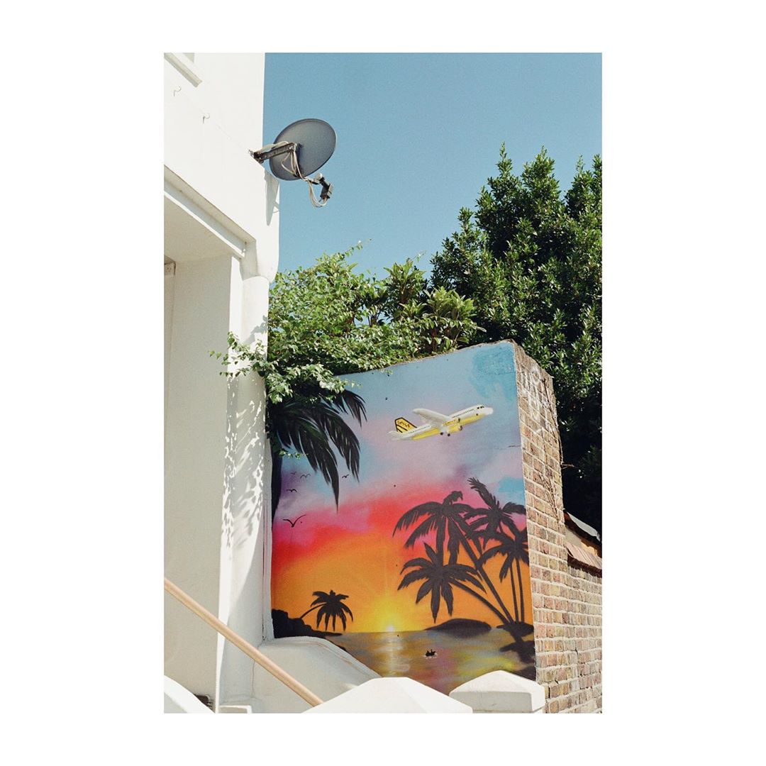 Mural of a tropical beach scene with palm trees and a plane on a brick wall under a clear sky, framed by a white building.