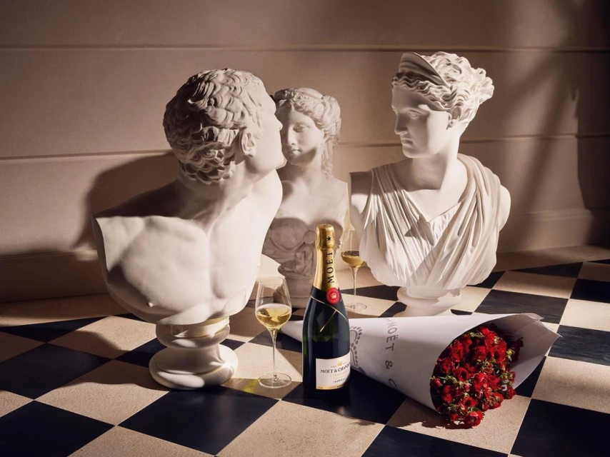Still life featuring classical bust sculptures, a champagne bottle, glasses of champagne, and a bouquet of red roses on checkered floor.