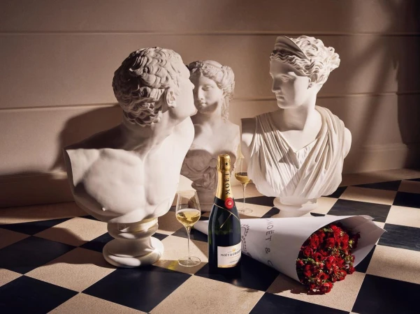 Still life featuring classical bust sculptures, a champagne bottle, glasses of champagne, and a bouquet of red roses on checkered floor.