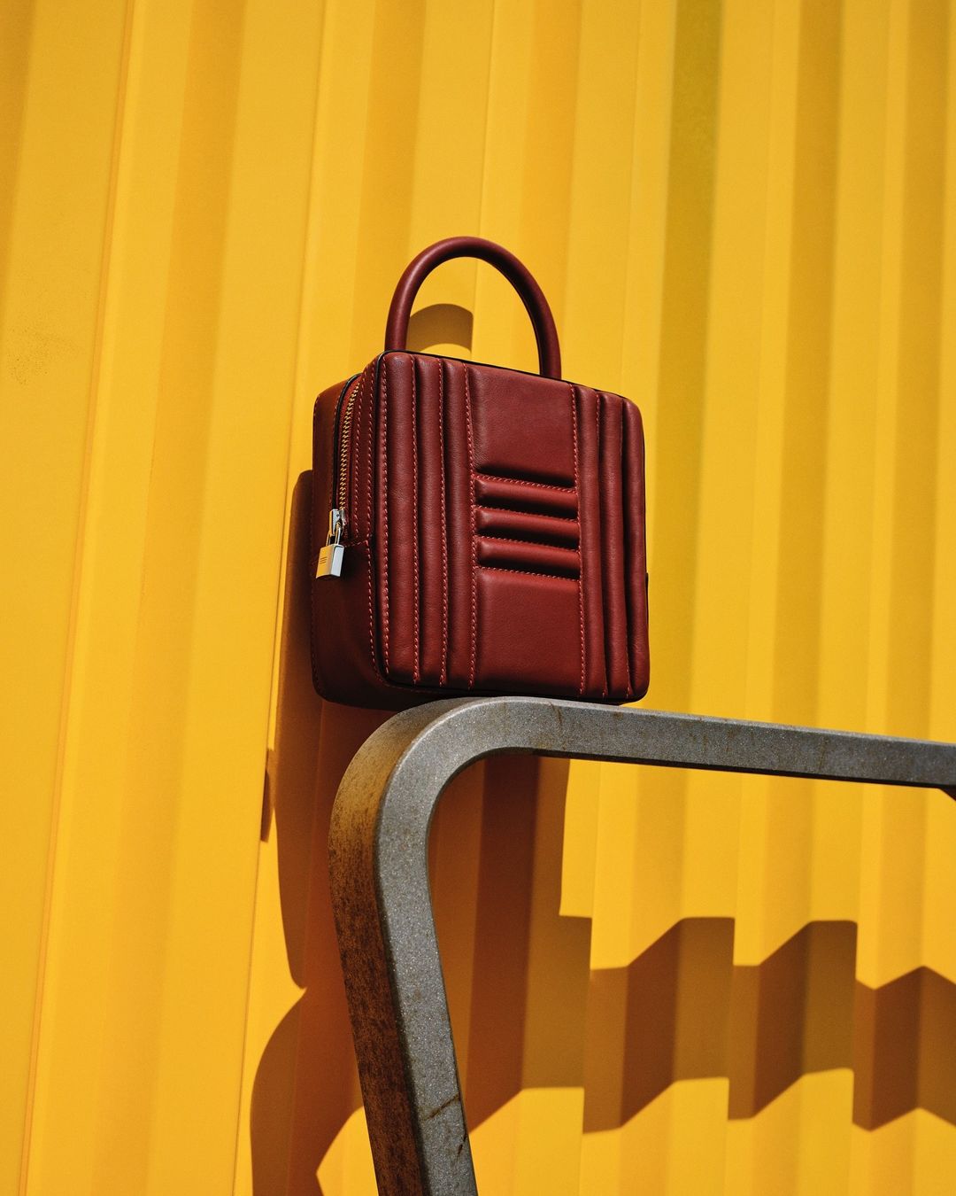 Burgundy leather handbag with a top handle, presented against a vibrant yellow corrugated metal wall. The scene is bathed in sunlight.