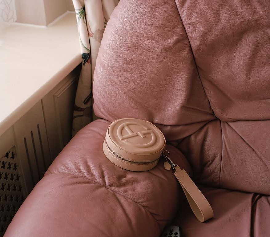 Close-up of a round, light brown cosmetic travel case with a monogram and short strap, sitting on a tufted, pink-toned leather couch.