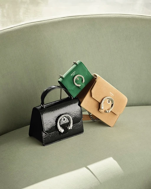 Three Jimmy Choo handbags in black patent leather, emerald green, and tan suede are stacked against a textured green wall in a product shot.