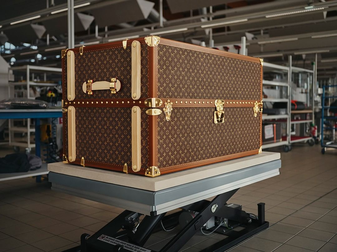 Luxury Louis Vuitton trunk, featuring a monogram pattern and gold accents, sits on a grey platform in a factory setting with metal racks.