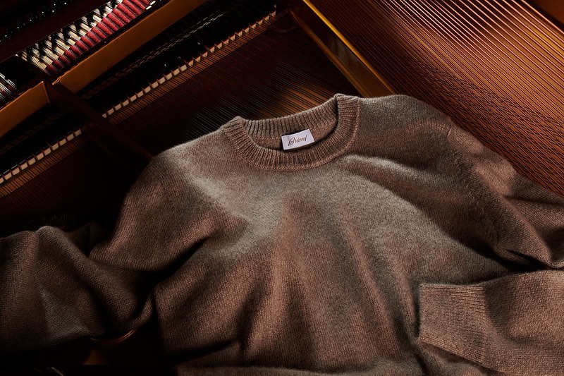 Elegant wool sweater with a visible 'Brioni' label, presented against a background of dark piano strings creating a luxurious feel.