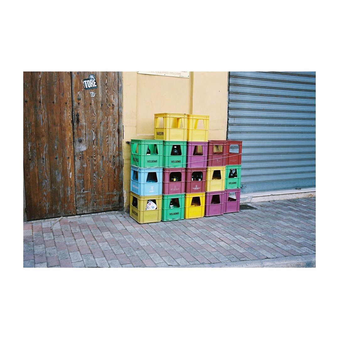 Colorful plastic crates stacked against a building with a wooden door and a metal garage door, set on a brick sidewalk.