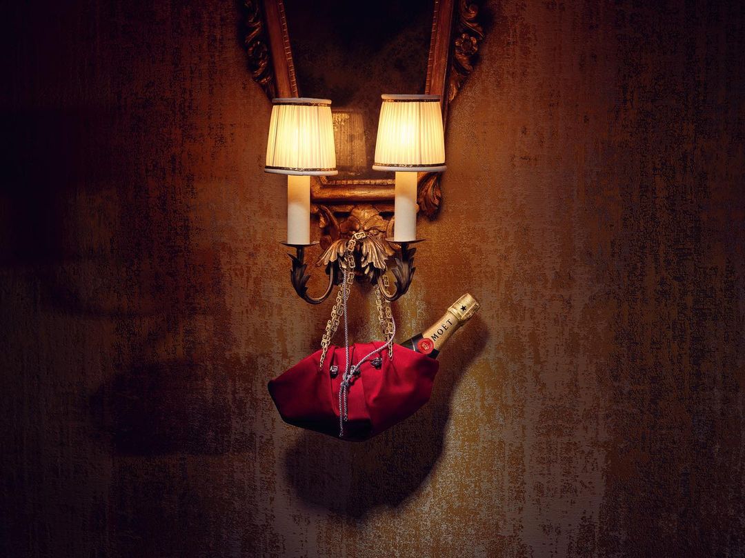 Bottle of Moet champagne in red fabric holder hanging from sconce with lamps against patterned wallpaper in moody lighting.