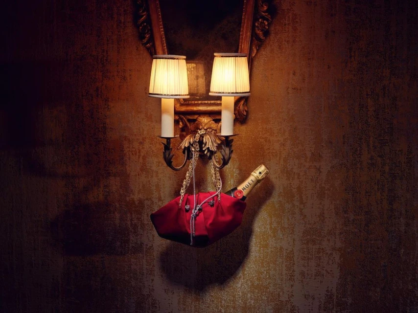 Bottle of Moet champagne in red fabric holder hanging from sconce with lamps against patterned wallpaper in moody lighting.