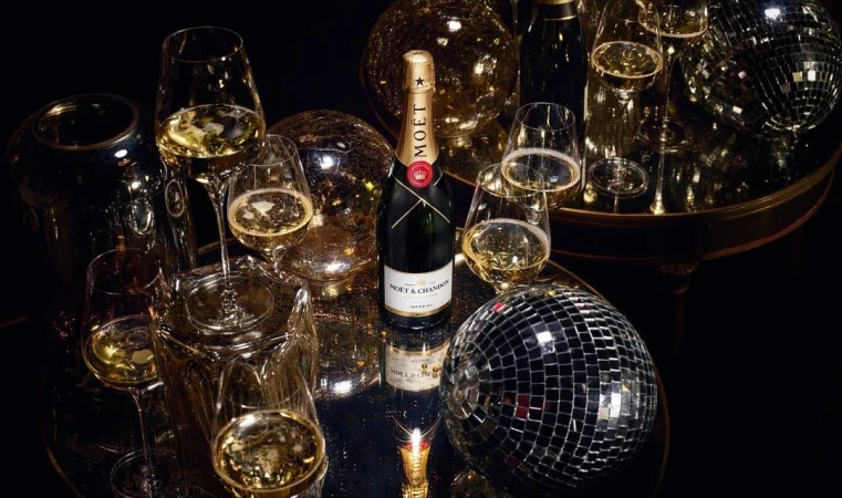 Moët & Chandon champagne bottle on mirrored surface surrounded by champagne flutes and disco balls reflecting golden light.