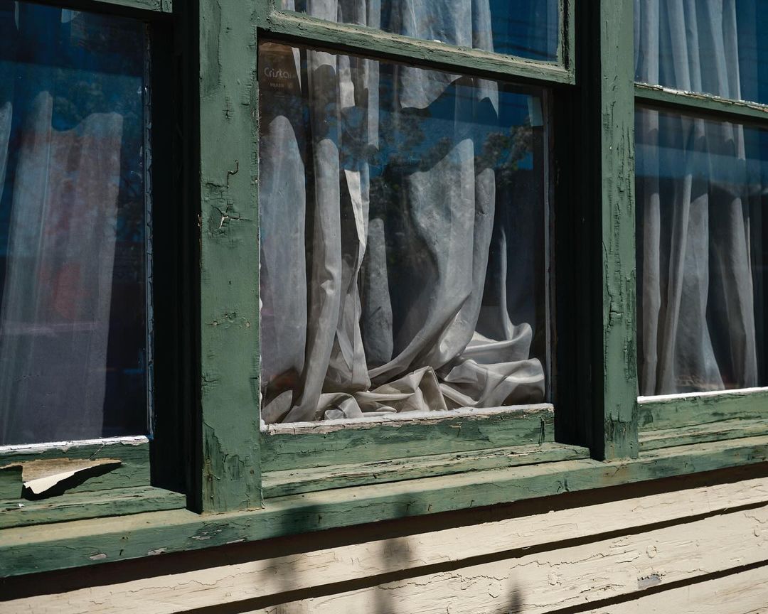 Close-up on an old window with peeling green paint and draped curtains. The window reflects outdoor light and hints at age.