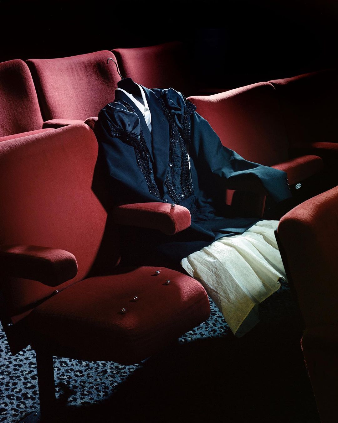 Formal dress and skirt on red theater seats, spotlight and shadow contrast, creating elegant fashion scene in an unexpected setting.