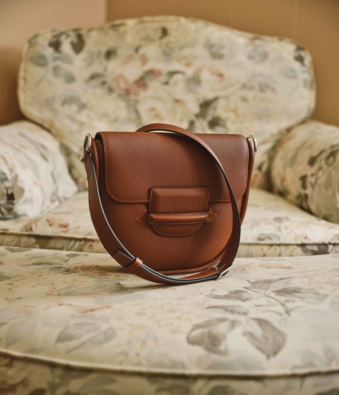 Brown leather saddle bag with flap closure on a floral armchair, illuminated by soft, diffused light, conveying luxury.