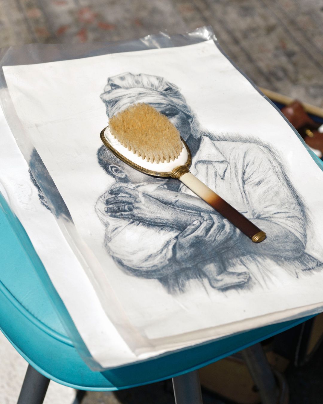 Pencil drawing of a woman and child with a hairbrush placed on top, set on a blue surface, captured in an indoor setting.