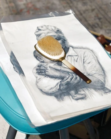 Pencil drawing of a woman and child with a hairbrush placed on top, set on a blue surface, captured in an indoor setting.