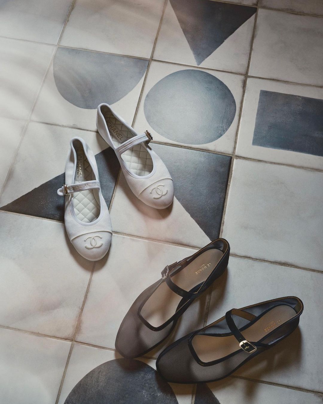 Two pairs of Chanel ballet flats, one white and one black, are arranged diagonally on a gray and white patterned tile floor.