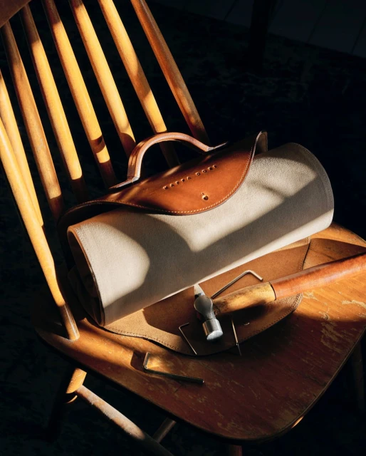 A handcrafted canvas and leather bag sits on a wooden chair in warm lighting, showcasing artisan craftsmanship and natural materials.