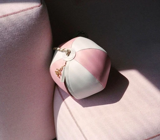 Stylish round handbag featuring a pink and white color block design, a top handle, and gold accents displayed on a pink velvet surface.